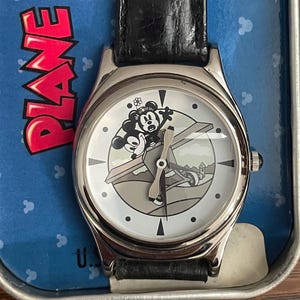 May include: A silver wristwatch with a black leather strap. The watch face depicts Mickey and Minnie Mouse in a biplane. The watch is presented in a blue tin, with the word "PLANE" in red.