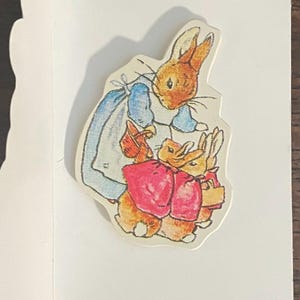 May include: A die-cut sticker featuring a watercolor illustration of a mother rabbit in a blue dress and two smaller rabbits in red jackets. The rabbits are depicted in a classic storybook style, with soft colors and detailed linework.
