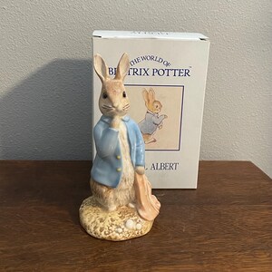 Puede incluir: Una figurita de porcelana de Peter Rabbit de Beatrix Potter, de la historia de Peter Rabbit. La figurita lleva una chaqueta azul y sostiene una zanahoria. La figurita está de pie sobre una base con un diseño marrón y amarillo. La caja de la figurita está en el fondo y dice "The World of Beatrix Potter" y "Albert".