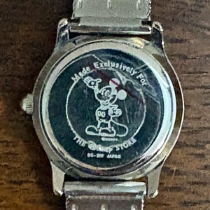 May include: Silver-toned wristwatch with a mesh band. The watch face features a black background with a Mickey Mouse graphic and the text "Made Exclusively For The Disney Store". The watch is marked "DS-329 JAPAN".