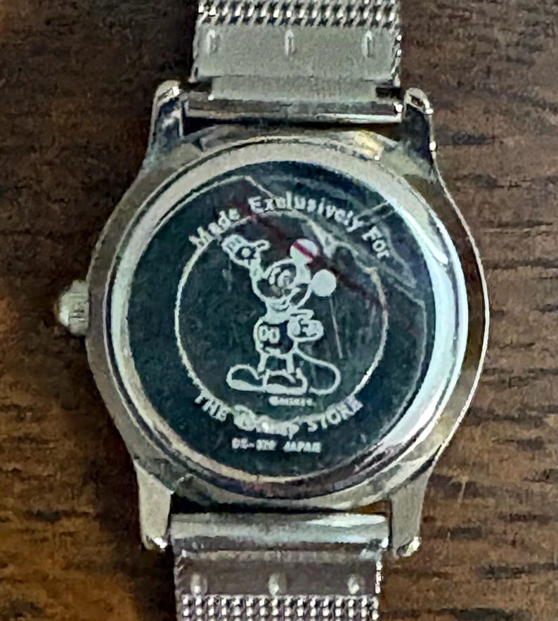 May include: Silver-toned wristwatch with a mesh band. The watch face features a black background with a Mickey Mouse graphic and the text "Made Exclusively For The Disney Store". The watch is marked "DS-329 JAPAN".
