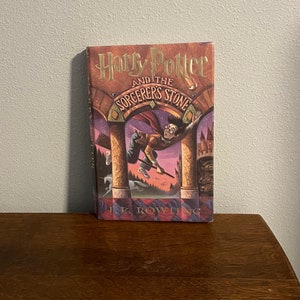 May include: A book cover for "Harry Potter and the Sorcerer's Stone" by J.K. Rowling. The cover features a purple and orange illustration of Harry Potter flying on a broomstick with a castle in the background.