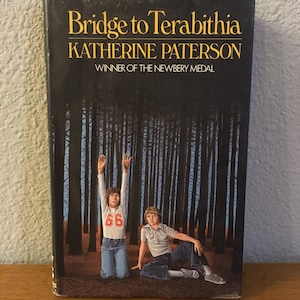 May include: A book cover with the title "Bridge to Terabithia" by Katherine Paterson, winner of the Newbery Medal. The cover features a dark forest scene with two children standing in the woods.