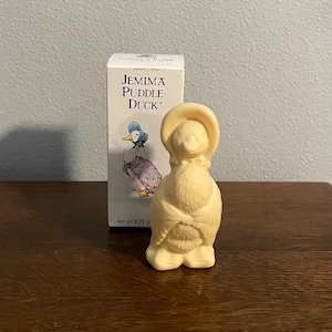 Puede incluir: Una figurita de jabón blanca de Jemima Puddle-Duck del libro infantil de Beatrix Potter. La figurita lleva un sombrero amarillo y un vestido blanco. El jabón está envasado en una caja blanca con las palabras "Jemima Puddle-Duck" impresas en ella. La caja también dice "peso neto 105 g".