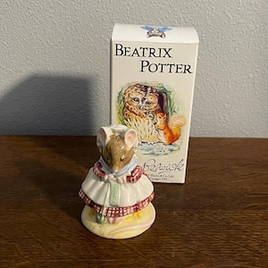 May include: A ceramic figurine of a mouse wearing a white dress with a red and pink trim, standing in front of a box with the text "Beatrix Potter". The box features illustrations of an owl and a squirrel.