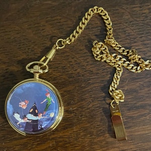 Op de afbeelding: Een gouden zakhorloge met een blauwe wijzerplaat met een scène uit Peter Pan. Het horloge heeft een gouden ketting met een gouden hanger.