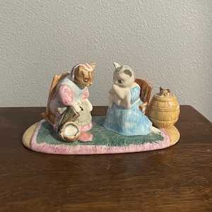 Puede incluir: Una figura de cerámica que representa a dos gatos con ropa de época sentados en una alfombra. Un gato lleva un vestido rosa y el otro un vestido azul. También hay un pequeño barril con un gato asomando. La figura está pintada en colores pastel.