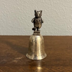 May include: A small, silver-toned bell with a cat figurine on top. The cat is wearing overalls and a vest. The bell is sitting on a wooden surface.