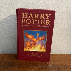 May include: A hardcover book titled "Harry Potter and the Order of the Phoenix." The cover is a deep red with gold lettering. A colorful illustration of a phoenix in flames is centered on the cover, with the author's signature below.