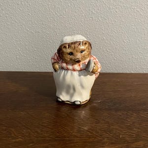 May include: A ceramic figurine of a hedgehog wearing a red and white striped dress and a white apron, holding a small iron. The figurine is brown, white, and red.