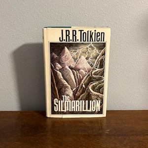 May include: A hardcover book titled The Silmarillion by J.R.R. Tolkien. The white dust jacket features a detailed illustration of jagged mountains under a lightning-filled sky, with the title and author name printed in bold black lettering.