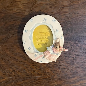 May include: A white oval picture frame with a blue and white floral design. The frame features a yellow background with the text "From the Tales of Beatrix Potter" and a small illustration of a rabbit. A small mouse figurine is attached to the frame, holding a red and white candy cane.