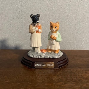 Puede incluir: Un conjunto de figuras de cerámica que representan un perro blanco y negro y un gato naranja que llevan delantales. El perro sostiene un libro y el gato sostiene un frasco. Las figuras están de pie sobre una base de piedra con una placa de madera que dice "Ginger and Pickles".