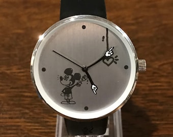 May include: A silver-toned wristwatch with a black leather band. The watch face features a cartoon Mickey Mouse character holding a paintbrush, with a heart design and pointing hands. The watch has a silver bezel and a crown.