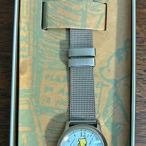 May include: A silver-toned wristwatch with a mesh band, presented in a decorative tin. The watch face features a light blue background with the image of a yellow bear. The watch hands point to the time.