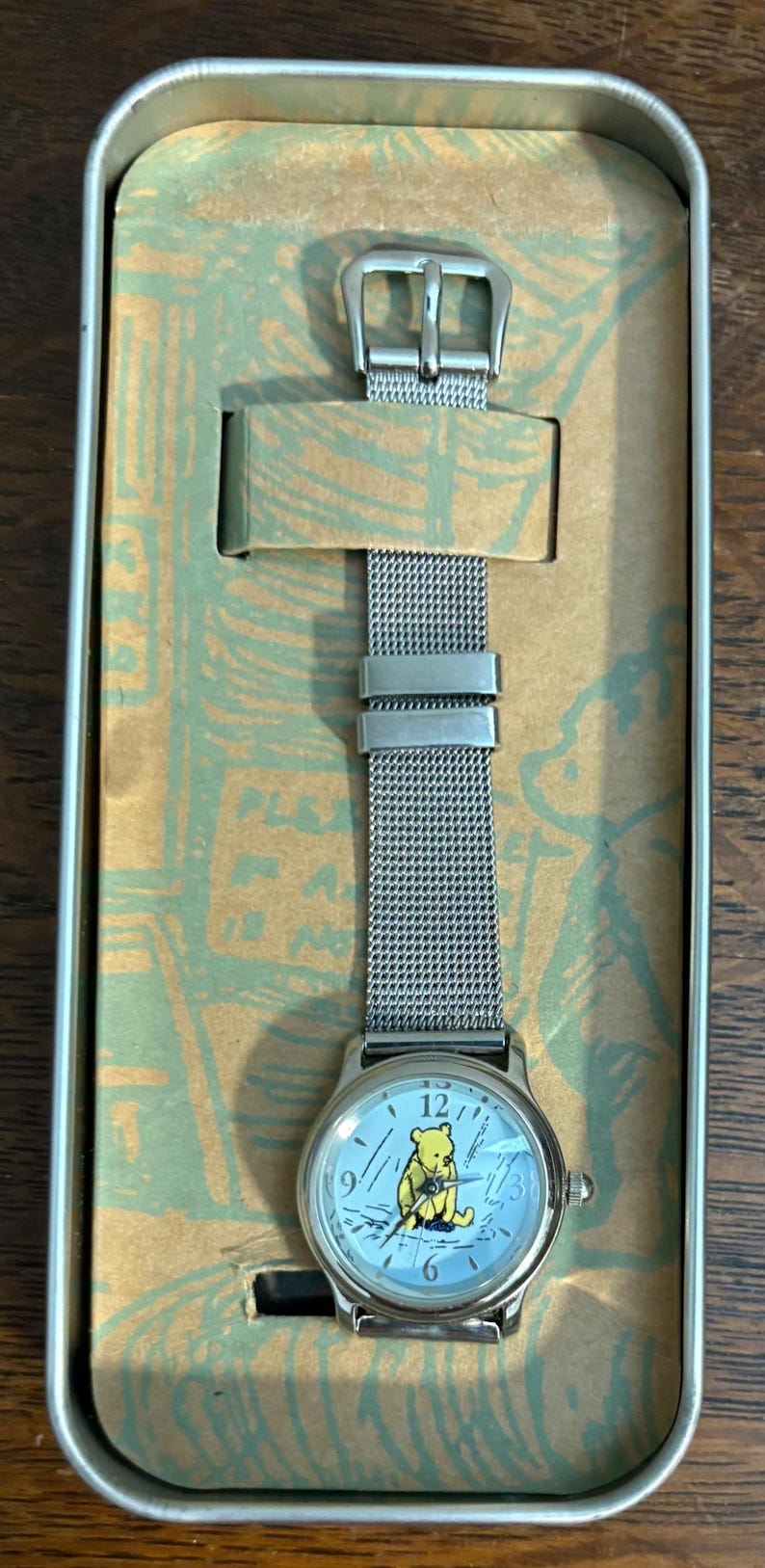 May include: A silver-toned wristwatch with a mesh band, presented in a decorative tin. The watch face features a light blue background with the image of a yellow bear. The watch hands point to the time.