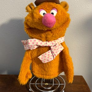May include: An orange plush bear puppet with a brown hat, pink nose, and a white bow tie with pink polka dots. The puppet is mounted on a metal stand, showcasing its design and features.