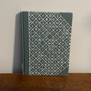 May include: A hardcover book with a dark teal cover featuring a white grid pattern filled with small, whimsical illustrations of plants, keys, and other objects. The book is standing upright on a wooden surface.