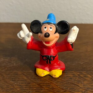 May include: A small figurine of Mickey Mouse in a sorcerer's outfit. The figure is wearing a red robe, yellow shoes, a blue hat, and white gloves. The figure is posed with arms outstretched and fingers pointing upwards.