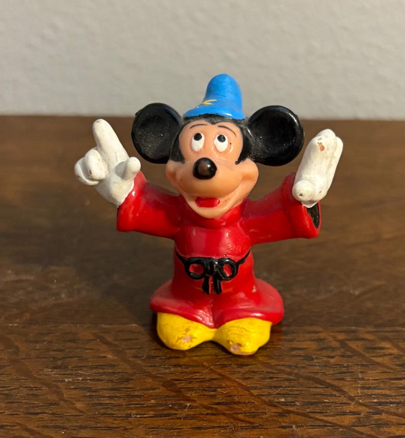 May include: A small figurine of Mickey Mouse in a sorcerer's outfit. The figure is wearing a red robe, yellow shoes, a blue hat, and white gloves. The figure is posed with arms outstretched and fingers pointing upwards.