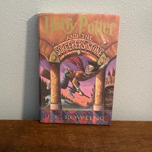 May include: A hardcover book titled "Harry Potter and the Sorcerer's Stone" with a colorful illustration of Harry Potter flying on a broomstick. The cover features a castle, a unicorn, and the author's name, J.K. Rowling.
