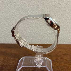 May include: A silver wristwatch with a mesh band is displayed on a clear plastic stand. The watch face is round with a silver bezel. The band is made of a fine, woven metal. The watch is presented at an angle, showcasing its design.