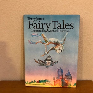 May include: A hardcover book titled "Fairy Tales" by Terry Jones, illustrated by Michael Foreman. The cover features whimsical illustrations of flying cat-like creatures, a castle, and a dragon. The book is set on a wooden surface.