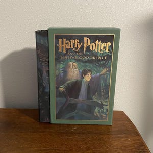May include: A green hardcover book with a gold title, "Harry Potter and the Half-Blood Prince". The cover features an illustration of Harry Potter and Dumbledore standing in a forest.