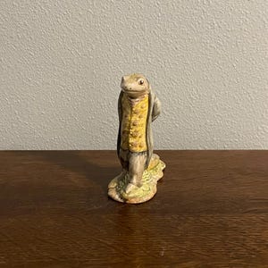 Può includere: Una statuetta in ceramica di una creatura simile a una lucertola che indossa un gilet giallo e un cappotto grigio. La statuetta ha un volto dettagliato e poggia su una base strutturata con sfumature verdi e gialle.