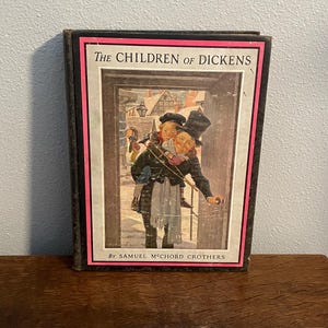 May include: A vintage book titled "The Children of Dickens" with a black cover and pink border. The cover illustration depicts a man carrying a child, with a snowy village scene in the background. The author's name, Samuel McChord Crothers, is also visible.