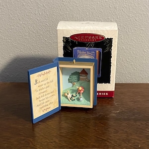Può includere: Un ornamento Hallmark Keepsake a forma di libro aperto. La pagina sinistra presenta il testo della filastrocca Jack and Jill. Il lato destro mostra una scena tridimensionale con una collina, un albero, un pozzo e due figure.