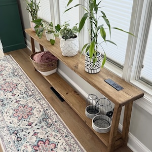 May include: A wooden console table with a shelf, featuring a variety of potted plants, including a large green plant in a black and white patterned pot. The table is placed in front of a window with white blinds. A patterned rug is on the floor in front of the table.
