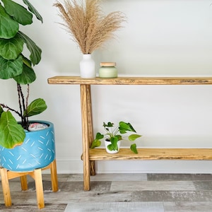 May include: A wooden console table with a light brown finish and a lower shelf. The table has a plant on the shelf and a vase with dried flowers on the top shelf.