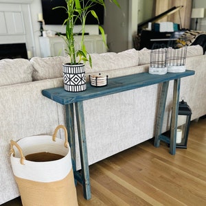 May include: A blue wooden console table with a white and black patterned planter, a set of coasters, and a black lantern. The table is in front of a beige sofa and has a distressed finish.