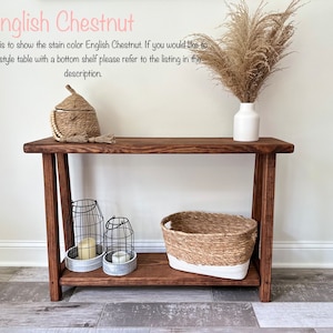 Puede incluir: Una mesa consola de madera con un estante inferior en color casta&ntilde;o ingl&eacute;s. La mesa tiene una cesta tejida, dos candelabros de alambre y una caja tejida con tapa en la parte superior. La mesa est&aacute; frente a una pared blanca con un jarr&oacute;n de hierba de la pampa seca.