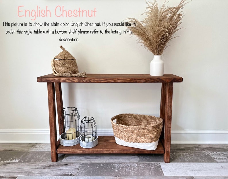Puede incluir: Una mesa consola de madera con un estante inferior en color casta&ntilde;o ingl&eacute;s. La mesa tiene una cesta tejida, dos candelabros de alambre y una caja tejida con tapa en la parte superior. La mesa est&aacute; frente a una pared blanca con un jarr&oacute;n de hierba de la pampa seca.