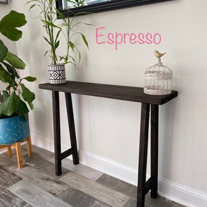 Puede incluir: Una mesa consola de madera marr&oacute;n oscuro con una maceta de patr&oacute;n blanco y negro en la parte superior. La mesa tiene un dise&ntilde;o simple en forma de A y est&aacute; etiquetada como "Espresso".