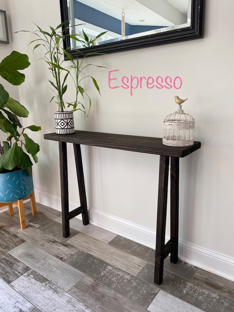 Puede incluir: Una mesa consola de madera marr&oacute;n oscuro con una maceta de patr&oacute;n blanco y negro en la parte superior. La mesa tiene un dise&ntilde;o simple en forma de A y est&aacute; etiquetada como "Espresso".