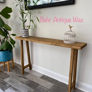 Puede incluir: Una mesa consola de madera con un acabado marr&oacute;n desgastado. La mesa tiene un dise&ntilde;o sencillo con dos patas en forma de A y una tapa rectangular. La mesa est&aacute; decorada con una planta en maceta y una jaula para p&aacute;jaros con una peque&ntilde;a figura de p&aacute;jaro. El texto "Behr Antique Wax" est&aacute; escrito en la pared detr&aacute;s de la mesa.