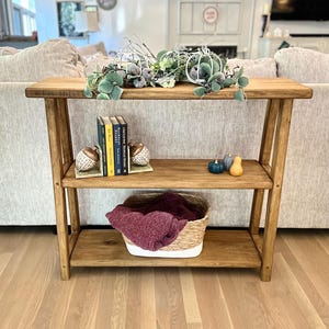May include: A wooden shelf unit with three shelves. The top shelf has a decorative arrangement of fall foliage and gourds. The middle shelf has a set of books with bookends. The bottom shelf has a woven basket with a burgundy blanket inside.