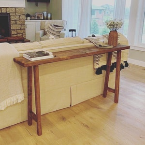 Puede incluir: Una mesa consola de madera r&uacute;stica con un acabado marr&oacute;n y veta de madera natural. La mesa tiene una tapa larga y rectangular y dos patas en forma de A. La mesa se coloca detr&aacute;s de un sof&aacute; beige y tiene un libro y un jarr&oacute;n con flores blancas en la parte superior.