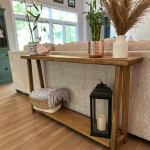 May include: A wooden console table with a lower shelf. The table has a natural wood finish and is decorated with a white ceramic vase, a potted plant in a copper pot, a potted plant in a white and black patterned pot, a white bird figurine, and a black lantern with a white candle.