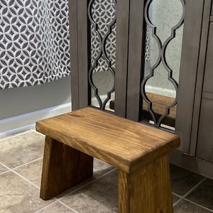 以下が含まれることがあります： 長方形の天板と角度のついた脚を持つ、濃い茶色の木製ステップスツール。スツールはバスルームのタイル張りの床の上に置かれています