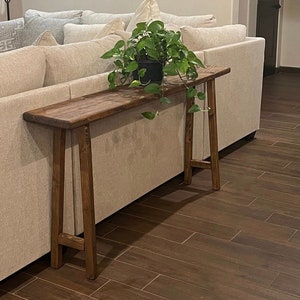 Puede incluir: Una mesa consola de madera marr&oacute;n oscuro con una superficie estrecha y dos patas en forma de A. La mesa se coloca junto a un sof&aacute; blanco y tiene una planta verde en maceta en la parte superior.