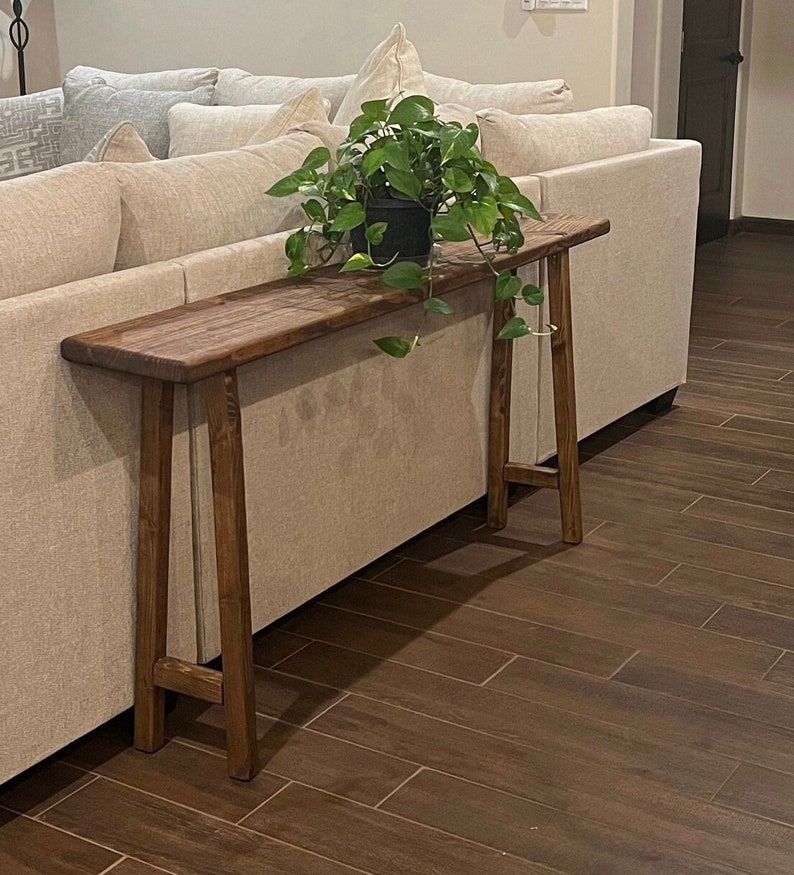 Puede incluir: Una mesa consola de madera marr&oacute;n oscuro con una superficie estrecha y dos patas en forma de A. La mesa se coloca junto a un sof&aacute; blanco y tiene una planta verde en maceta en la parte superior.