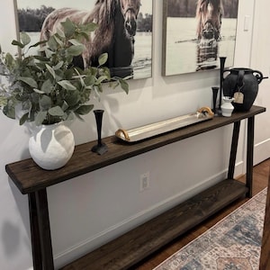 May include: A dark brown wooden console table with a lower shelf, displaying decorative items. A white textured vase holds greenery. A silver tray with wooden handles and black candlesticks are on the table. Two framed horse portraits hang on the wall.
