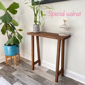 Puede incluir: Una mesa consola de madera con un tinte nogal oscuro. La mesa tiene un dise&ntilde;o simple y moderno con una tapa rectangular y dos patas en forma de A. La mesa est&aacute; decorada con una planta en maceta y una jaula para p&aacute;jaros con una figurita de p&aacute;jaro dorado.