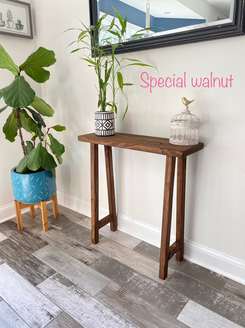 Puede incluir: Una mesa consola de madera con un tinte nogal oscuro. La mesa tiene un dise&ntilde;o simple y moderno con una tapa rectangular y dos patas en forma de A. La mesa est&aacute; decorada con una planta en maceta y una jaula para p&aacute;jaros con una figurita de p&aacute;jaro dorado.