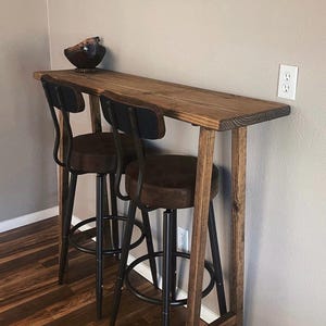 May include: A wooden bar table with two black metal bar stools. The table has a rustic wood finish and the stools have brown leather seats.