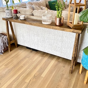 May include: A rustic wooden console table with a distressed finish. The table has a simple A-frame design and is placed in front of a white sofa. The table is decorated with a birdcage, potted plants, and a lantern.